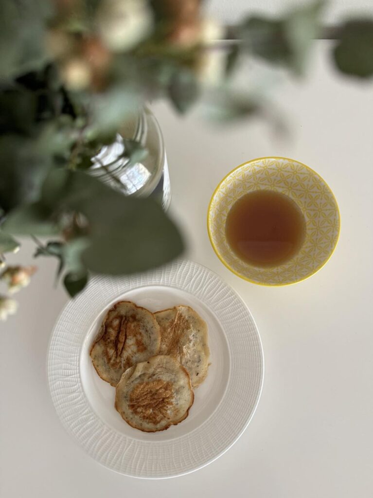 Eggless pancakes on a plate with a side of maple syrup and a vase of flowers in the top left corner of picture.