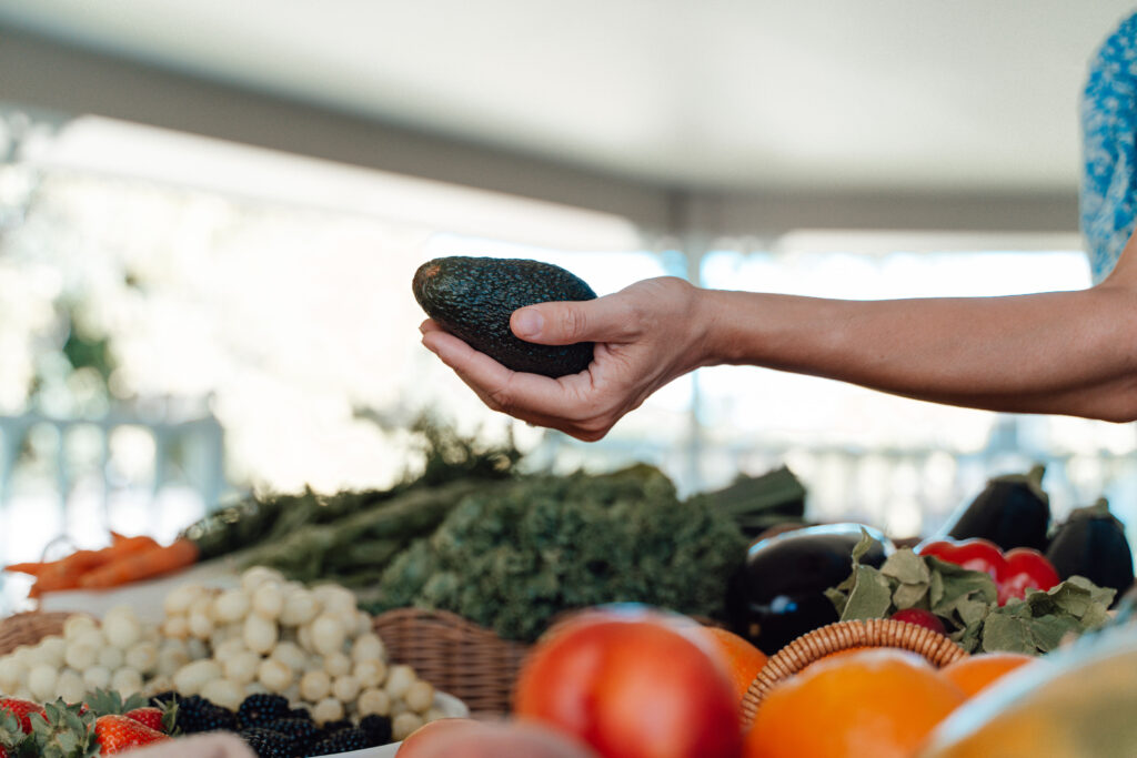 Woman holding avocado over a table of produce.