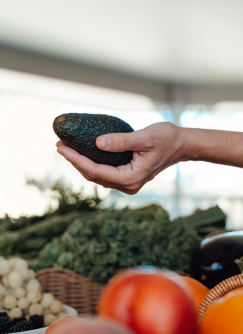 Woman holding avocado above a table of colourful produce.