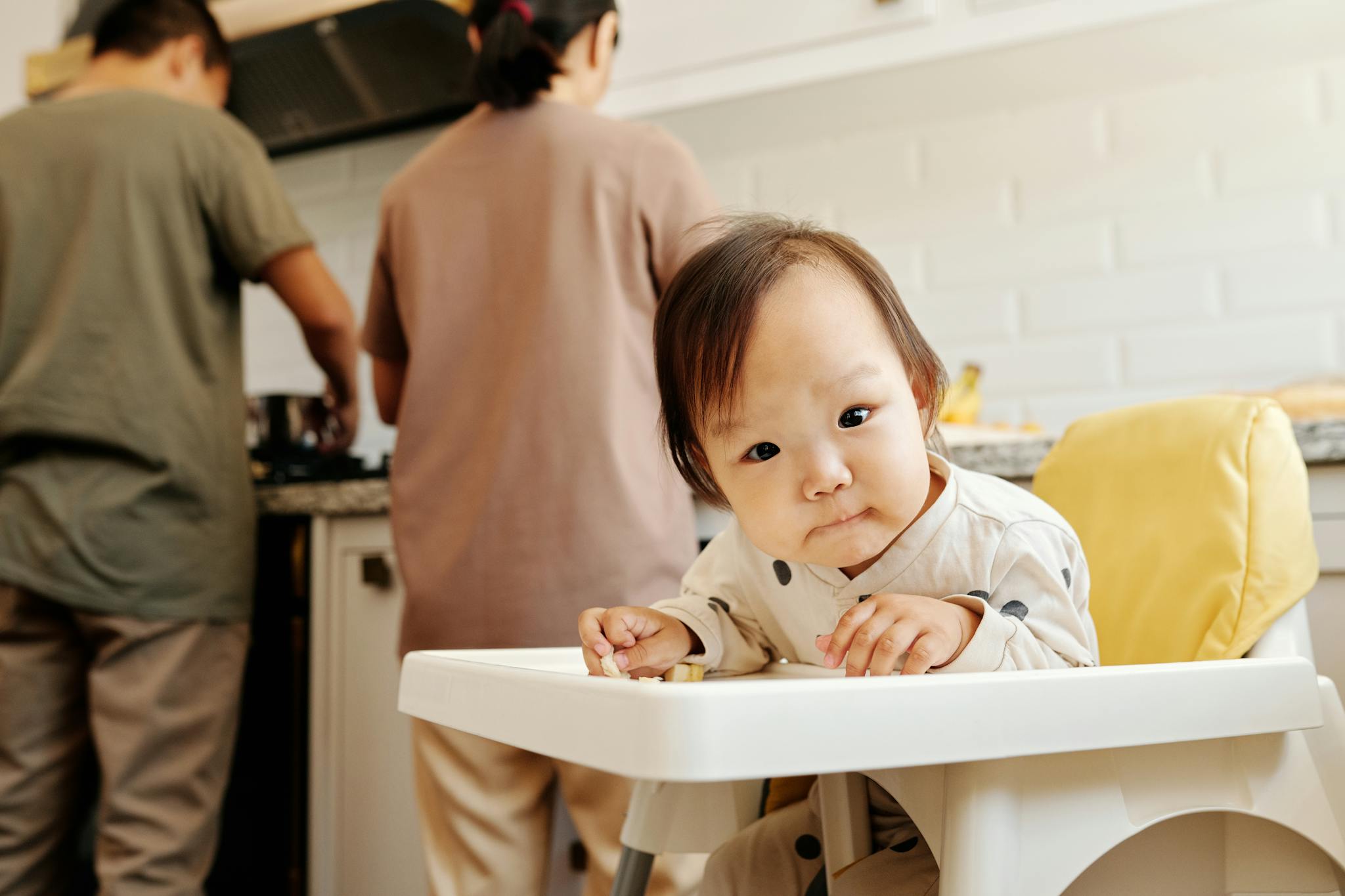 Cute toddler sitting in a high chair while parents prepare food in the kitchen.