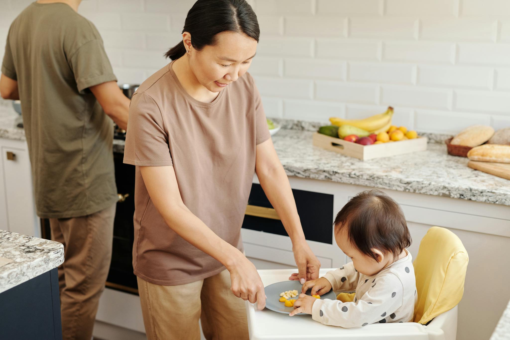 Mother feeding toddler in high chair with healthy fruits in a modern kitchen.